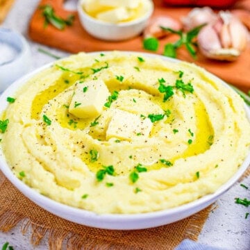 A white bowl of creamy mashed potatoes topped with butter and fresh parsley.