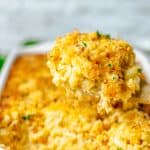 A wooden spoon scooping creamy Baked Mac and Cheese with cornbread crust out of a white baking dish.