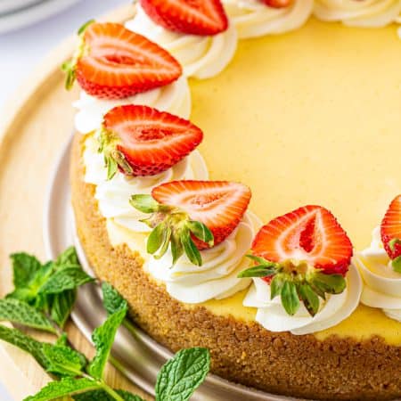 A whole vanilla bean cheesecake topped with whipped cream swirls and fresh strawberry halves.
