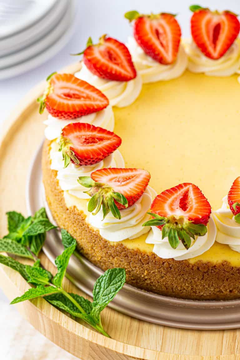 A whole vanilla bean cheesecake topped with whipped cream swirls and fresh strawberry halves.