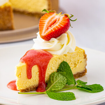 Slice of Vanilla Bean Cheesecake on white plate with garnishes and strawberry sauce.