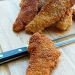 Golden crouton chicken strips piled on a wooden cutting board with a crispy crumb crust.