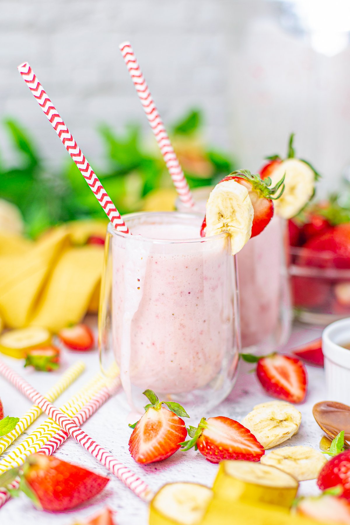 Two glasses of Strawberry Banana Smoothie with straws surrounded by fruit