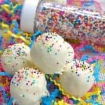 A pile of white cake truffles covered in rainbow sprinkles on festive party confetti with a bottle of sprinkles behind.