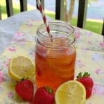 A mason jar of strawberry iced tea with a striped paper straw surrounded by fresh strawberries and lemon halves on a floral tablecloth.