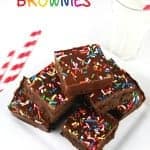 Fudgy black bean brownies topped with chocolate frosting and rainbow sprinkles stacked next to a glass of milk.