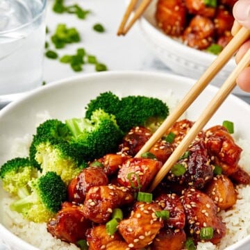 Glossy brown sugar glazed Chinese Chicken bites served over white rice with steamed broccoli, topped with sesame seeds and green onions in a white bowl.
