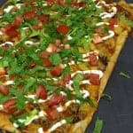 A taco pizza topped with seasoned beef, melted cheese, lettuce, diced tomatoes, and sour cream, cut into squares.