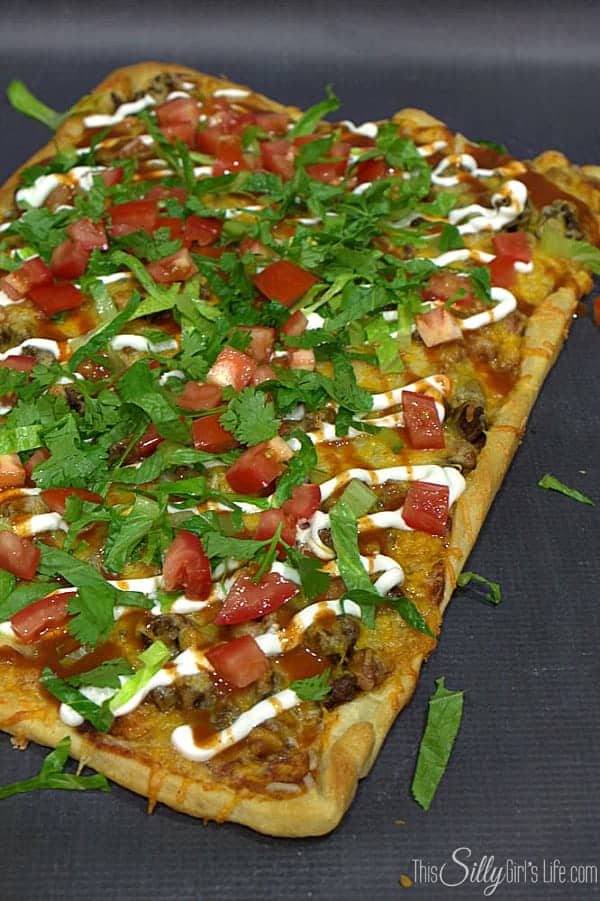 A taco pizza topped with seasoned beef, melted cheese, lettuce, diced tomatoes, and sour cream, cut into squares.