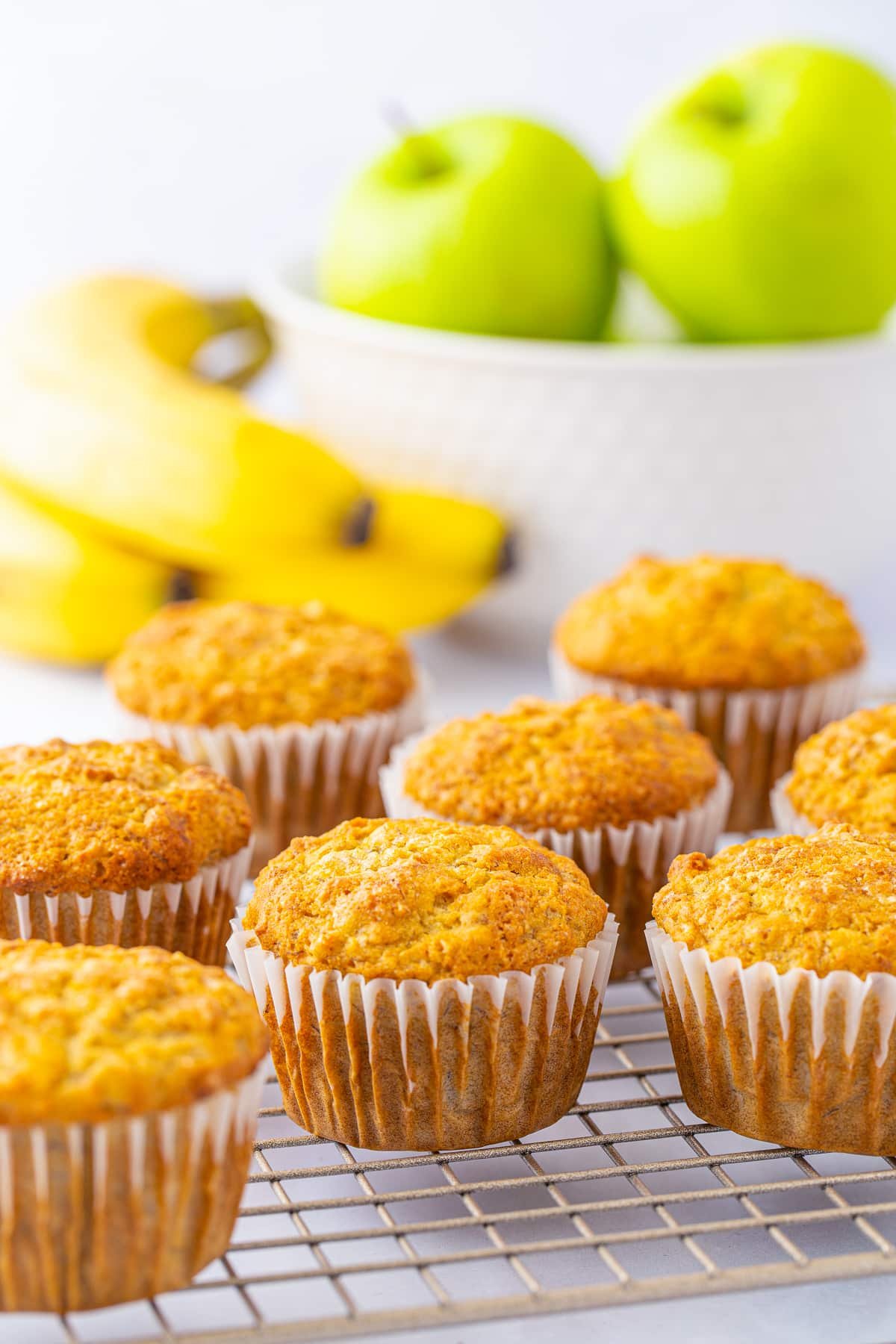 Apple Banana Oat Muffins on wire rack in liners with fruit in background.