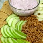 A glass bowl of pink cranberry dip on a wooden board surrounded by green apple slices, pretzels, and graham crackers.