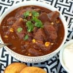 A white bowl of chunky Beef Chili topped with cilantro next to cornbread muffins and sour cream.