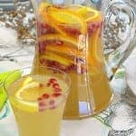 A glass pitcher of sparkling pomegranate punch loaded with orange slices, lemon slices, and pomegranate seeds beside a filled glass.