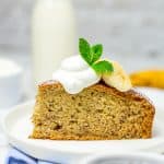 A slice of moist banana cake on a white plate dusted with powdered sugar and topped with fresh banana.