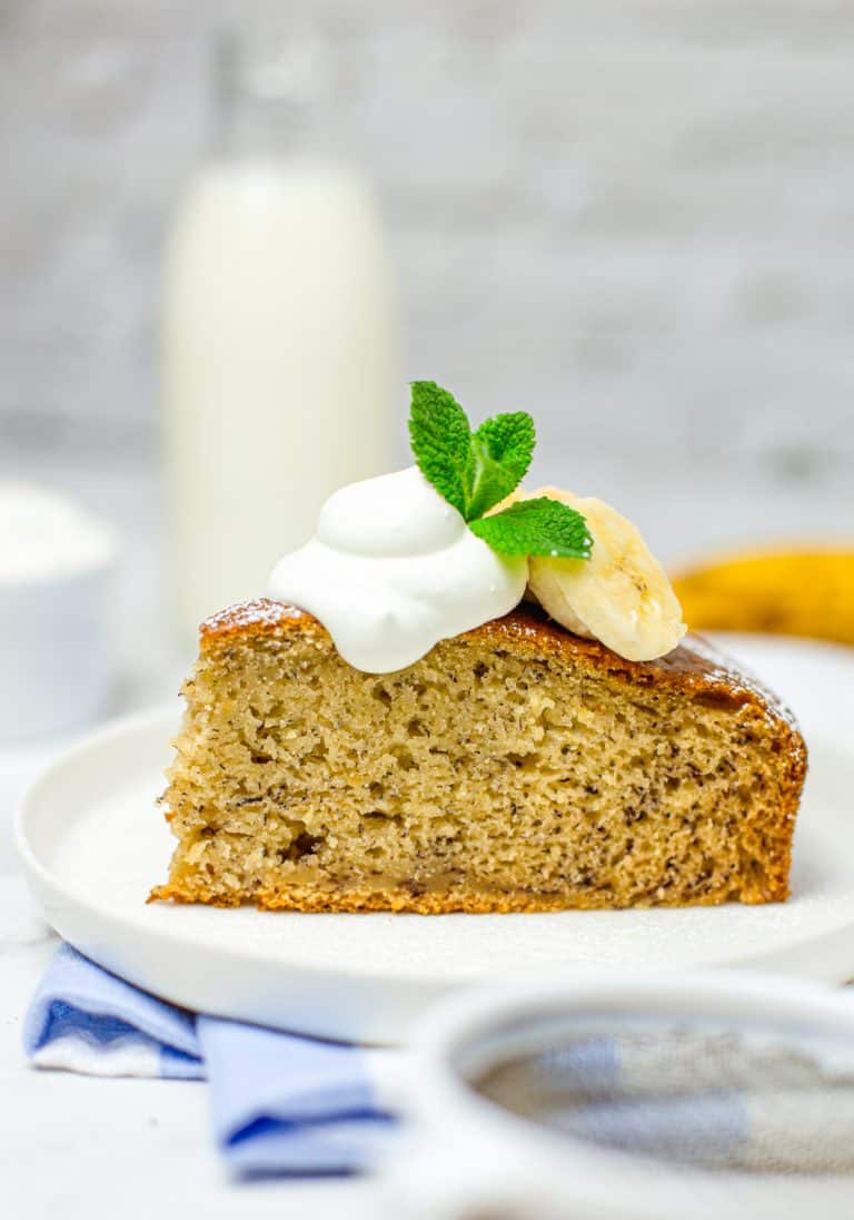 A slice of moist banana cake on a white plate dusted with powdered sugar and topped with fresh banana.