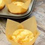 A cheese muffin in a parchment wrapper in front of a muffin tin full of golden muffins.