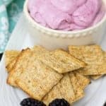 A white ramekin of purple marshmallow dip topped with a fresh blackberry surrounded by woven wheat crackers.