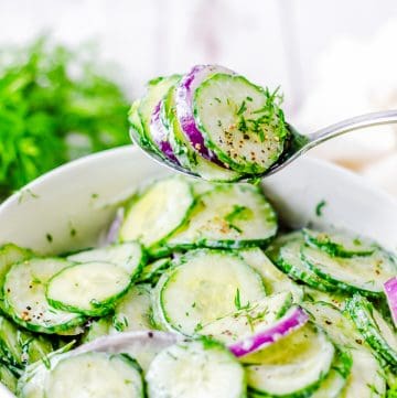 A spoon lifting a scoop of creamy Cucumber Dill Salad with red onion from a white bowl.