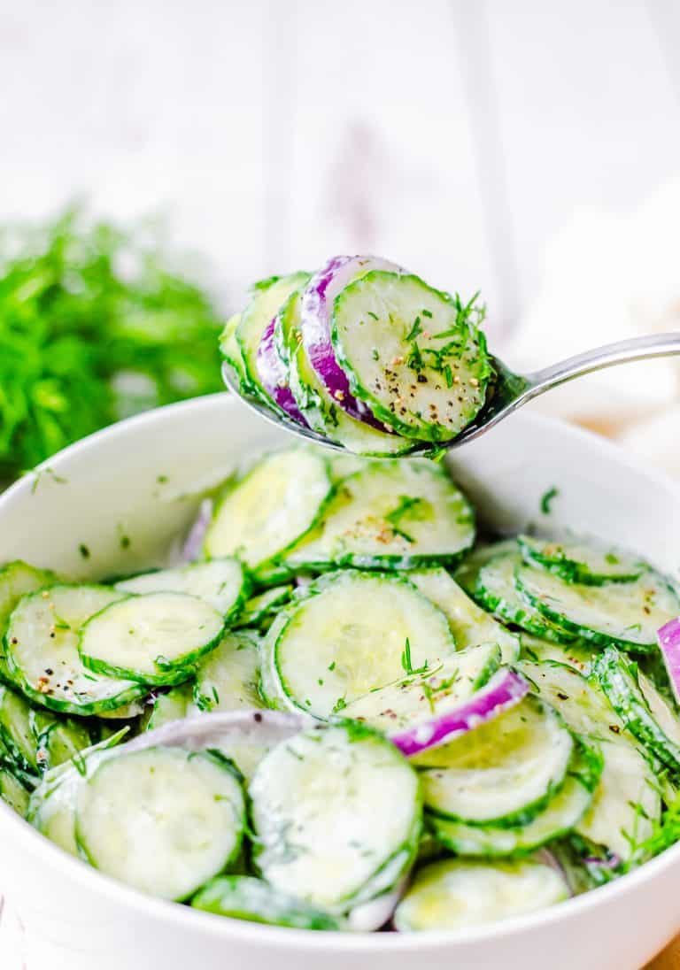 A spoon lifting a scoop of creamy Cucumber Dill Salad with red onion from a white bowl.