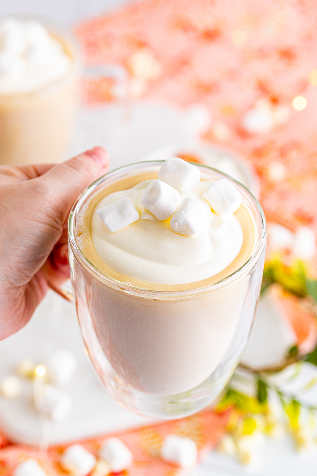 Hand holding up mug of Boozy White Hot Chocolate Recipe topped with whipped cream and marshmallows.