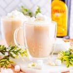 Two glass mugs of kahlua hot chocolate topped with whipped cream and marshmallows in front of a Kahlua bottle.