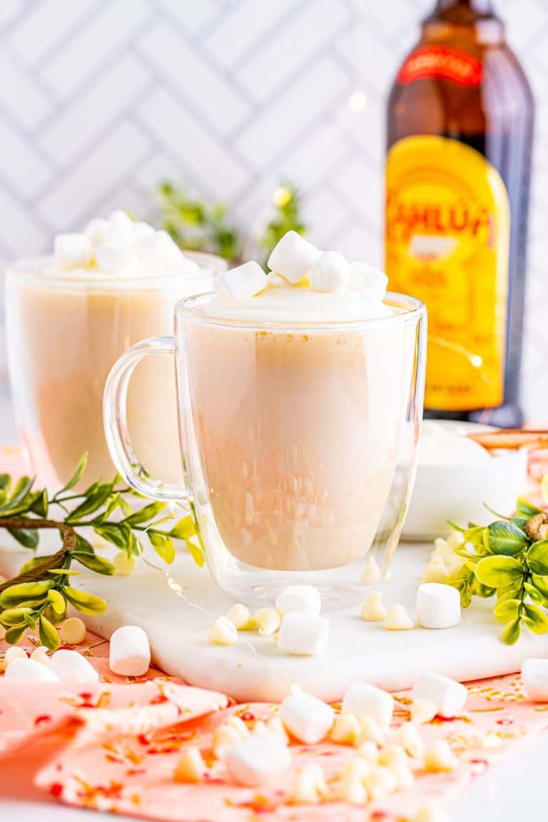 Two glass mugs of kahlua hot chocolate topped with whipped cream and marshmallows in front of a Kahlua bottle.