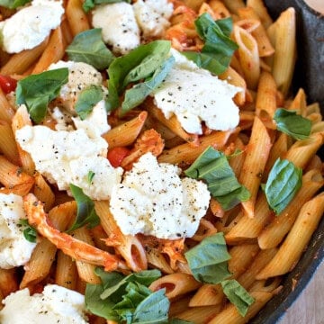 A cast iron skillet of creamy chicken fra diavolo pasta topped with melted ricotta and torn fresh basil.