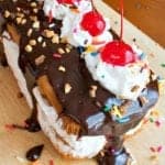 A hot fudge sundae cake loaf topped with fudge dripping down the sides, whipped cream, chopped nuts, sprinkles, and maraschino cherries.