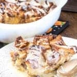A serving of eggnog cinnamon roll bread pudding with icing drizzle on a white plate in front of the baking dish.