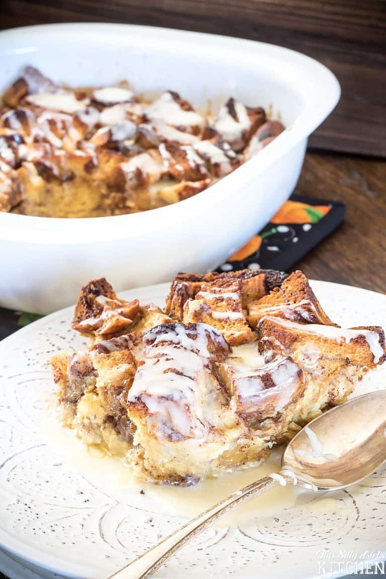 A serving of eggnog cinnamon roll bread pudding with icing drizzle on a white plate in front of the baking dish.