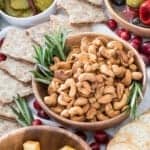 A wooden bowl of roasted cashews with fresh rosemary sprigs surrounded by crackers, cheese cubes, and grapes on a snack board.