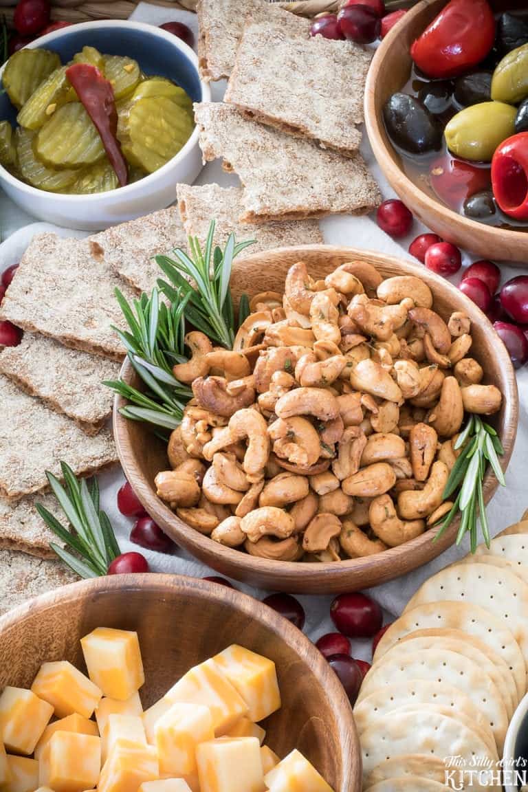 A wooden bowl of roasted cashews with fresh rosemary sprigs surrounded by crackers, cheese cubes, and grapes on a snack board.