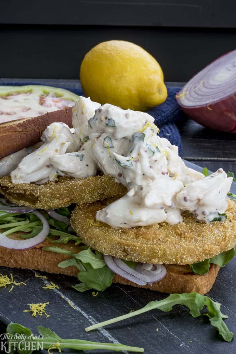 Fried green tomato sandwich stacked with crab salad, arugula, and red onion on toasted white bread.