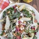 Overhead view of a warm kale salad in a white bowl topped with bacon, pear slices, goat cheese, red onion, and dried cranberries.
