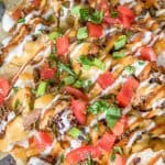 A sheet tray of loaded BBQ beef nachos with cheese sauce, tomatoes, and scallions.