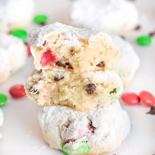 A stack of Snowball Cookies with the top cookie broken open showing chocolate chips and candy pieces inside.