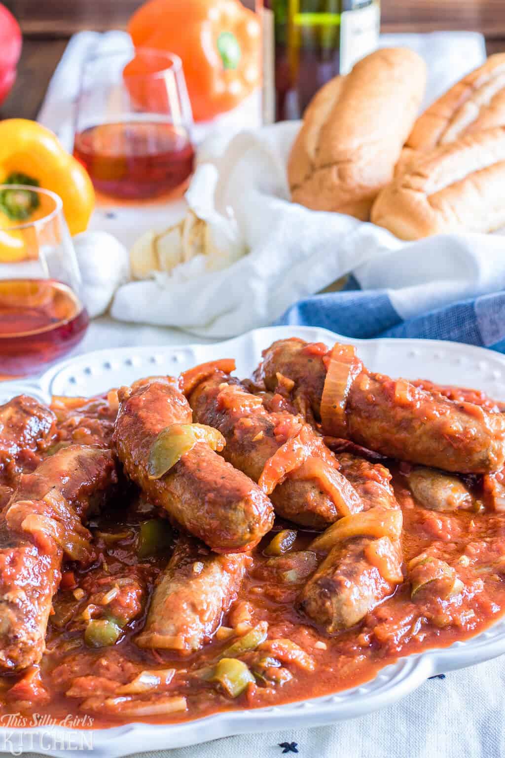 Marsala Sausage and Peppers on white platter with wine and rolls in background.