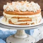 A naked coconut cake on a white stand topped with toasted coconut and a caramel drizzle.