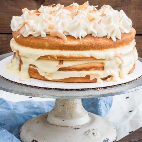 A naked coconut cake on a white stand topped with toasted coconut and a caramel drizzle.