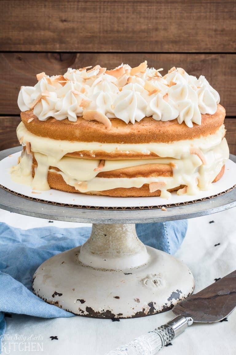 A naked coconut cake on a white stand topped with toasted coconut and a caramel drizzle.