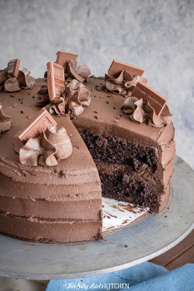 A whole chocolate layer cake with whipped chocolate frosting and chocolate shavings, one slice cut to show the moist layers.