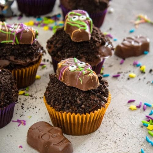 Halloween Cupcakes topped with cookie crumbs and candy splattered chocolate skulls on a gray counter.