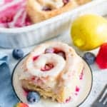 A glazed lemon sweet roll on a white plate with fresh blueberries, a strawberry, and a lemon.