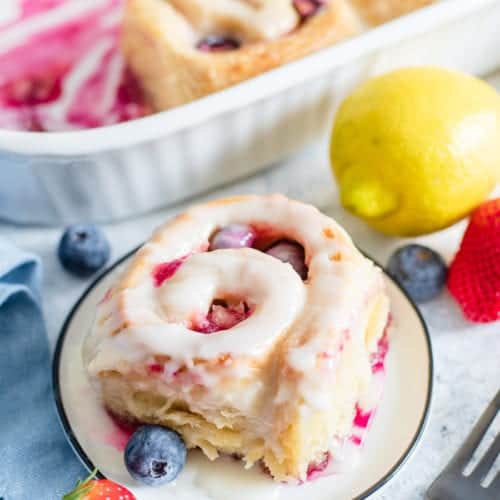 A glazed lemon sweet roll on a white plate with fresh blueberries, a strawberry, and a lemon.