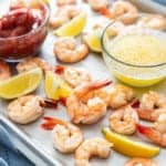 Roasted shrimp cocktail on a sheet tray with cocktail sauce, drawn butter, and lemon wedges.