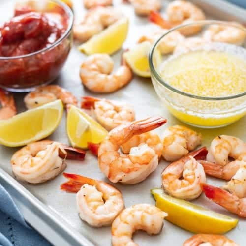 Roasted shrimp cocktail on a sheet tray with cocktail sauce, drawn butter, and lemon wedges.