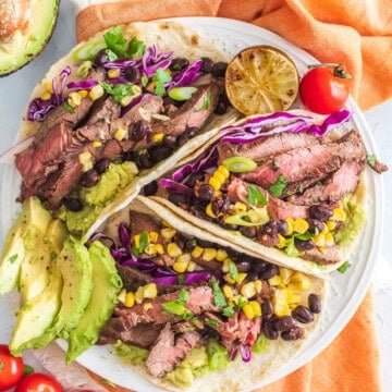 A plate of skirt steak tacos with avocado, corn, cabbage, and lime.