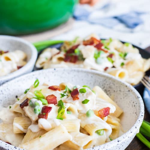 A bowl of creamy Ranch Chicken Pasta topped with crispy bacon and scallions with a green dutch oven behind it.
