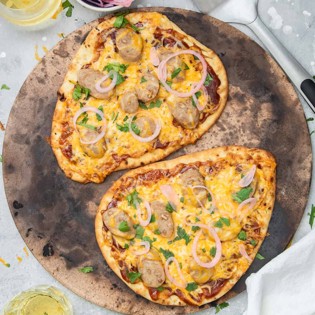 Two BBQ chicken pizzas on naan topped with cheese, chicken sausage, cilantro, and pickled onions.
