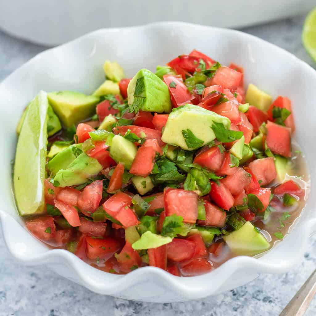 Overhead view of avocado salsa in a white bowl served with lime and tortilla chips.